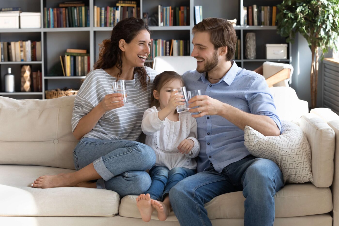 A Smiling mother, father, and young daughter sit together on a living room couch, each holding a glass of water and enjoying a drink.